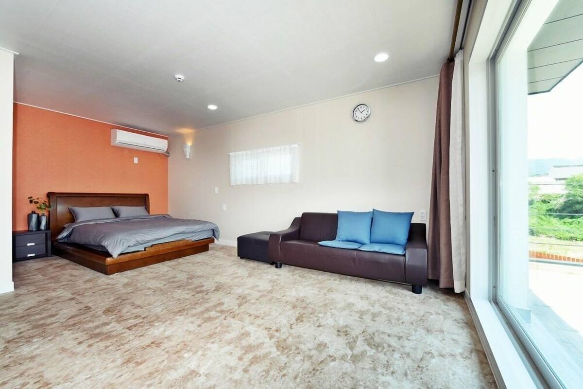 A bedroom with a bed, a sofa, an air conditioning unit, a clock, and a window with curtains.