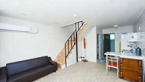 Una sala de estar con un sofá oscuro, una escalera, una pequeña cocina con un lavamanos y armarios, y una puerta que da a otra habitación.