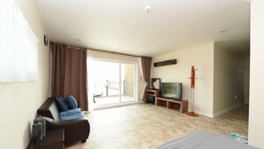 Una habitación amplia con un gran ventanal corredero que da a un balcón, un sofá de color oscuro con cojines azules, un televisor de pantalla plana sobre un mueble de madera y una pequeña mesita de noche de madera.