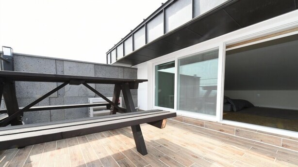 Un patio al aire libre con una mesa y bancos de madera, un muro de concreto y un amplio ventanal corredizo que da a un espacio interior.