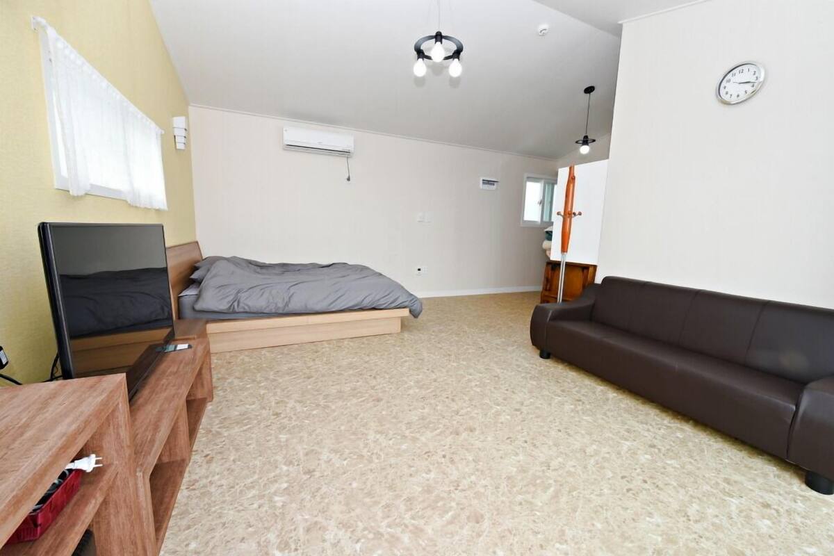 A bedroom with a bed, a wooden nightstand, a brown leather sofa, a ceiling light, and a wall clock.