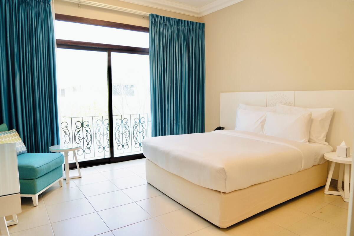 A hotel room with a large bed, a small table, a sofa, and a balcony with curtains.