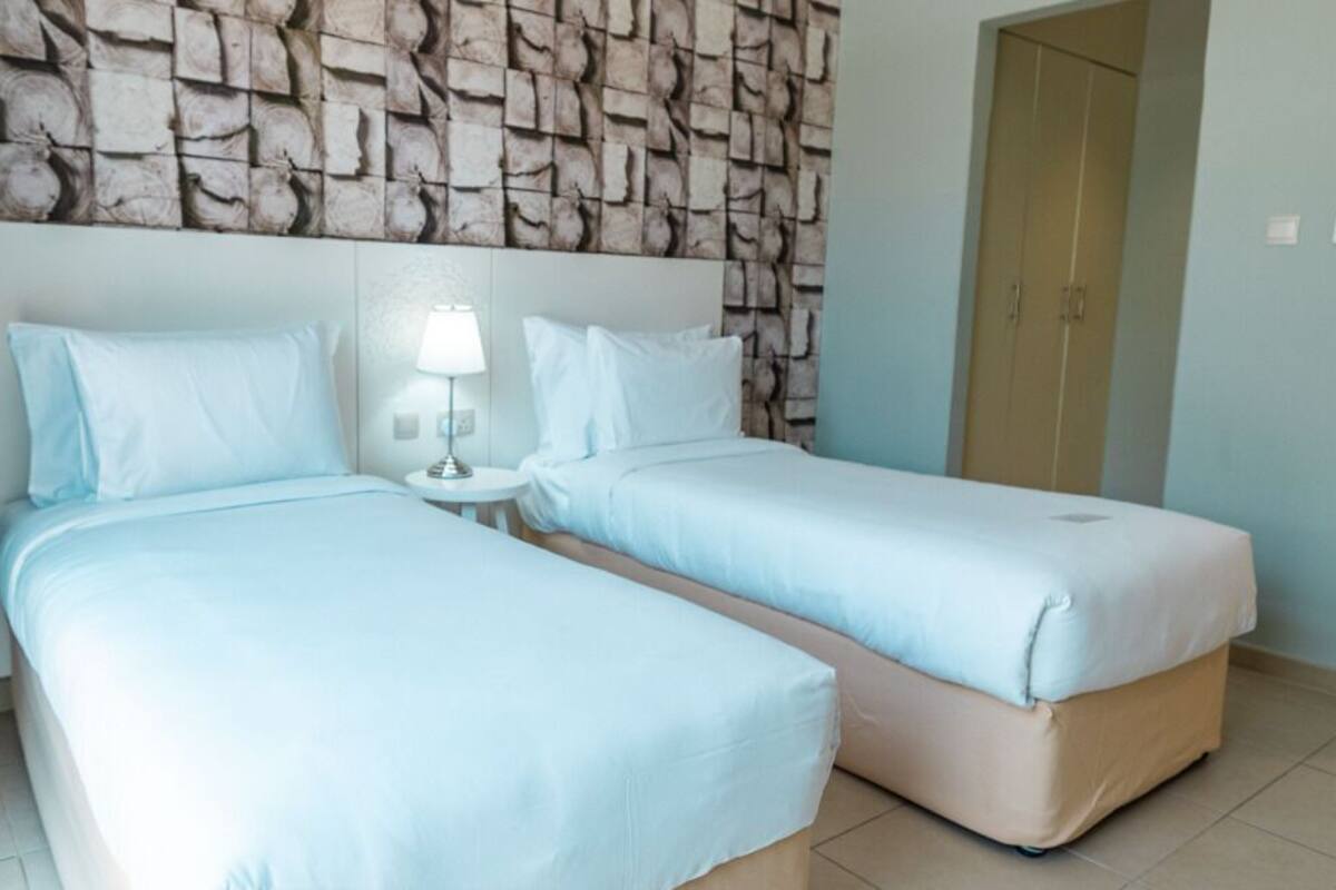 A hotel room with two beds, a stone-patterned wall, a wooden door, and a small bedside table with a lamp.
