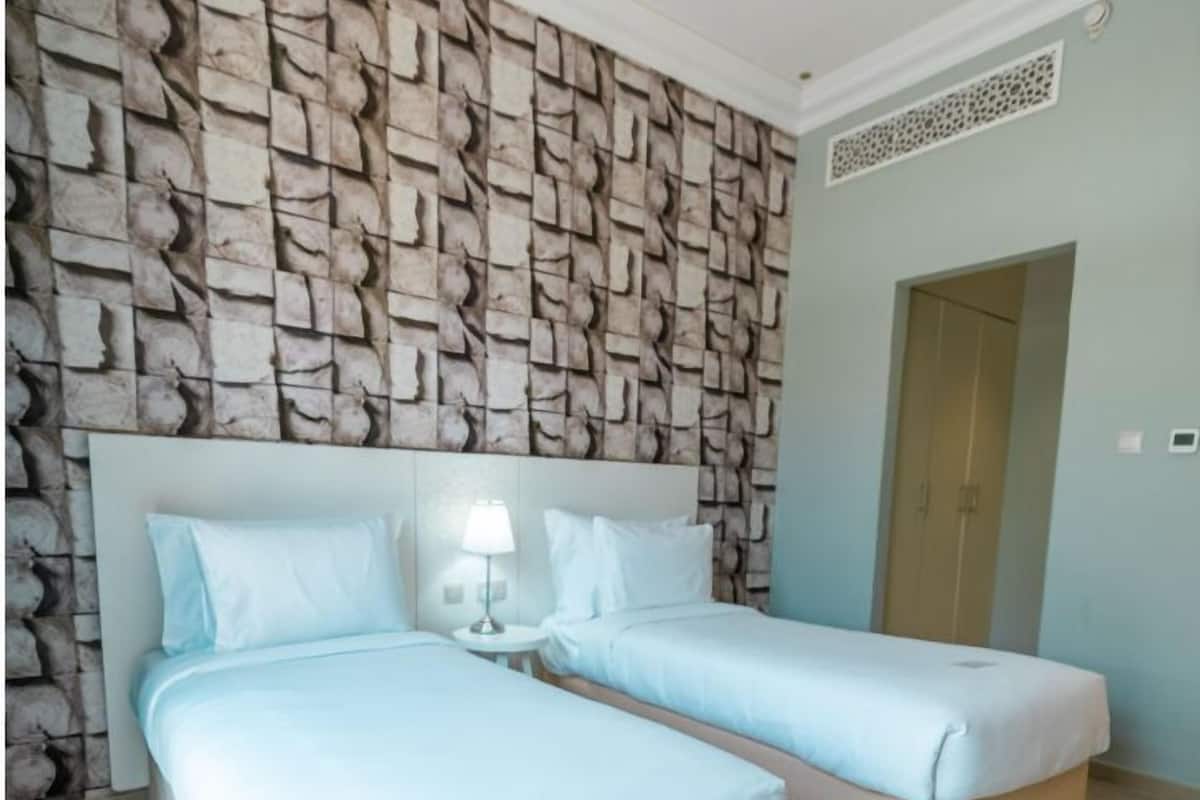 A hotel room with two beds, a stone-patterned wall, a small table with a lamp, and a door.