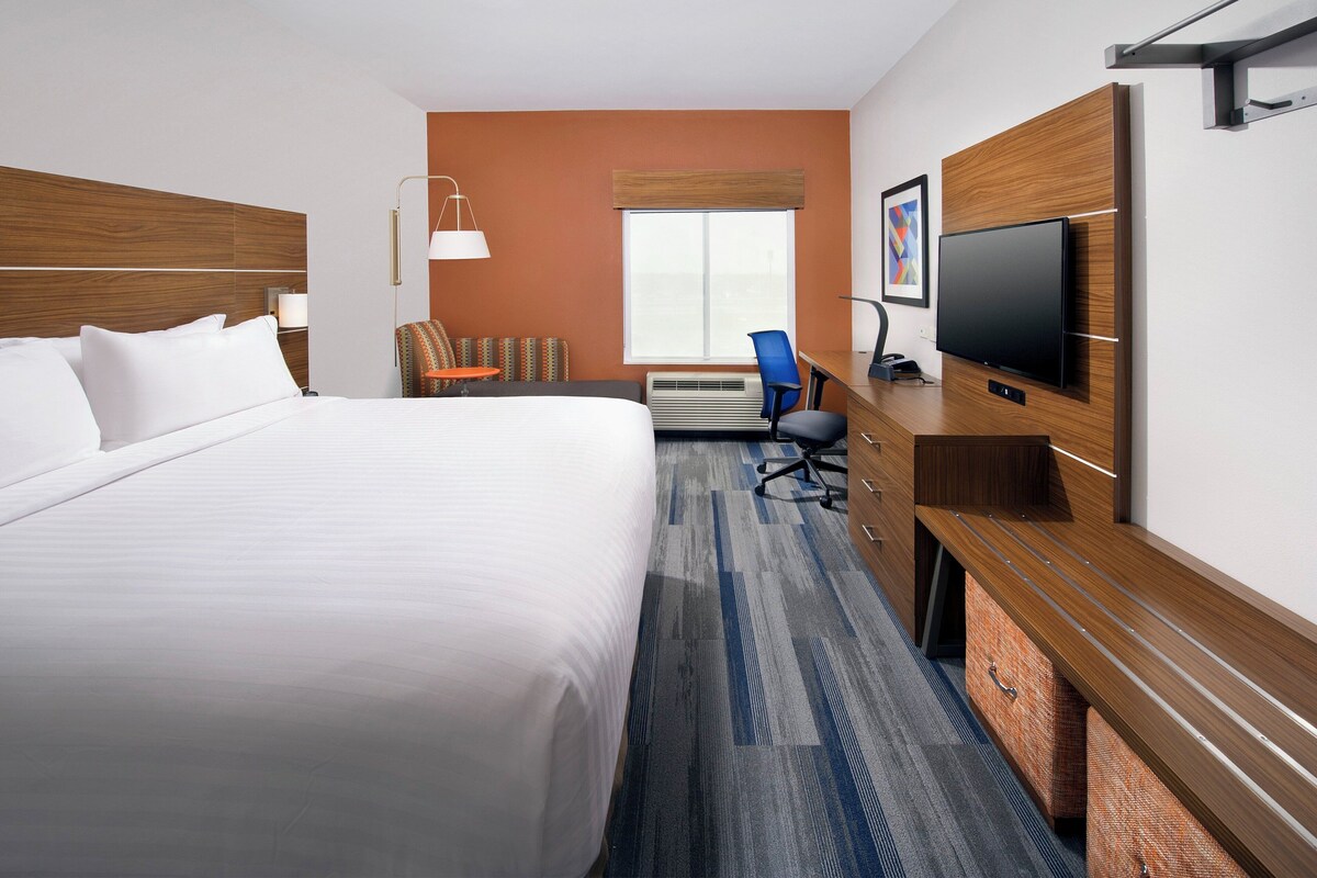A hotel room with a large bed, a desk with a flat-screen TV, a chair, and a window with blinds.