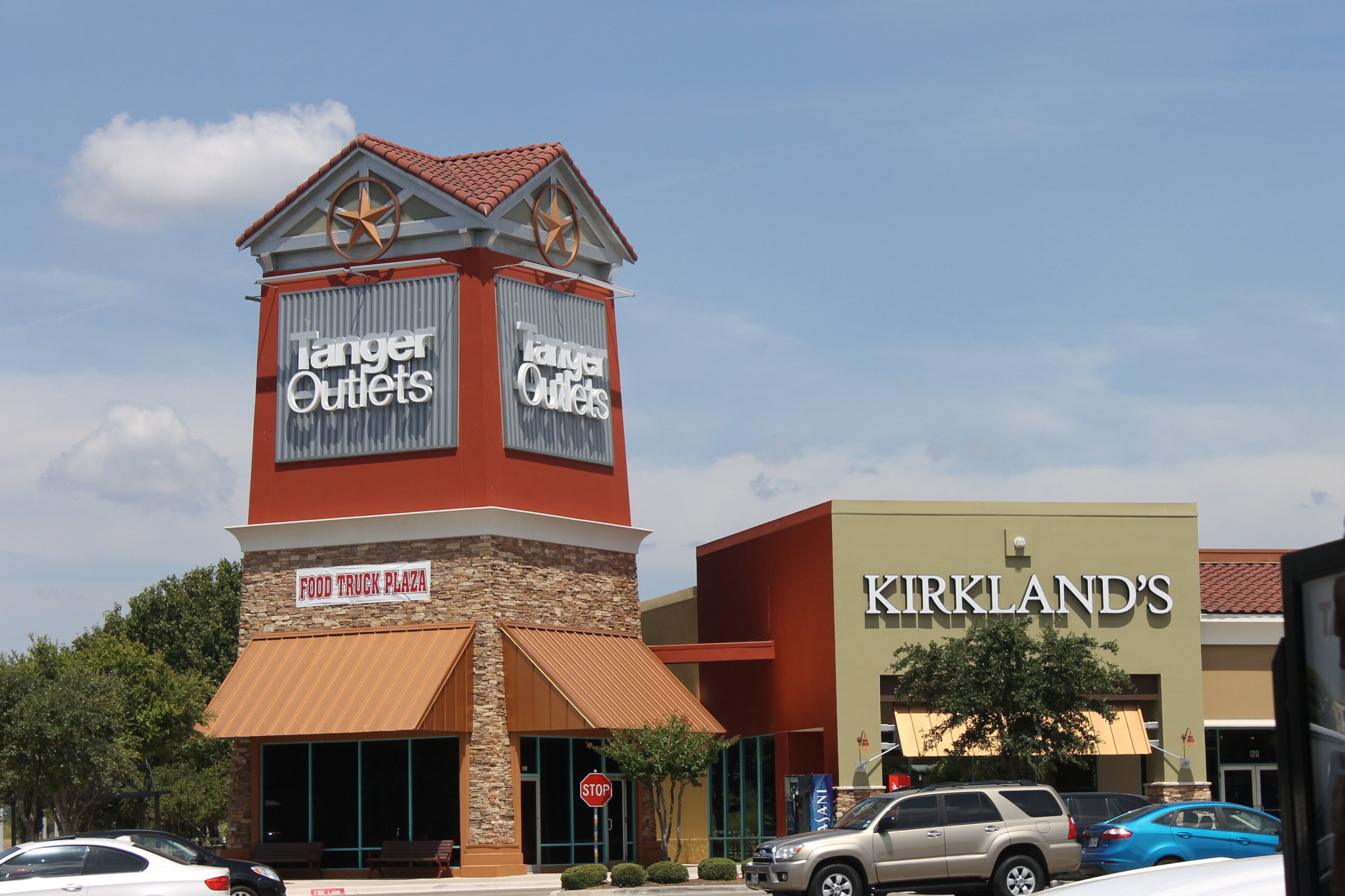 A commercial building with a tower featuring 'Tanger Outlets' signage, a 'Food Truck Plaza' sign, and 'Kirkland's' store.
