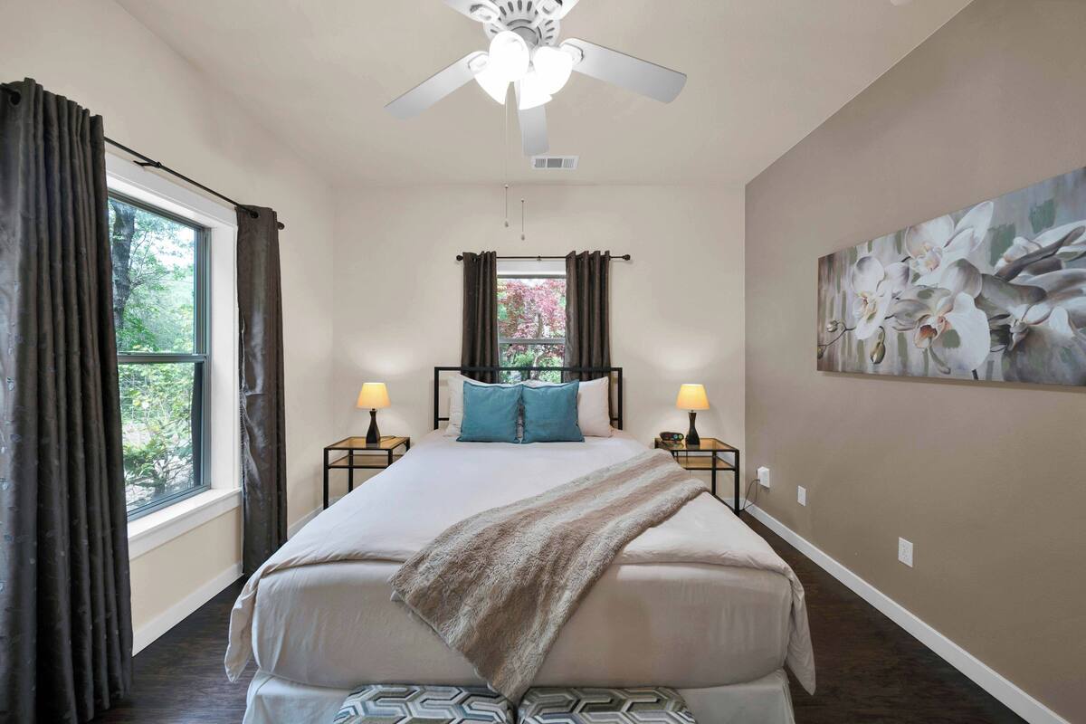 A bedroom with a large bed, a ceiling fan, two bedside lamps, a window with curtains, and a wall painting.