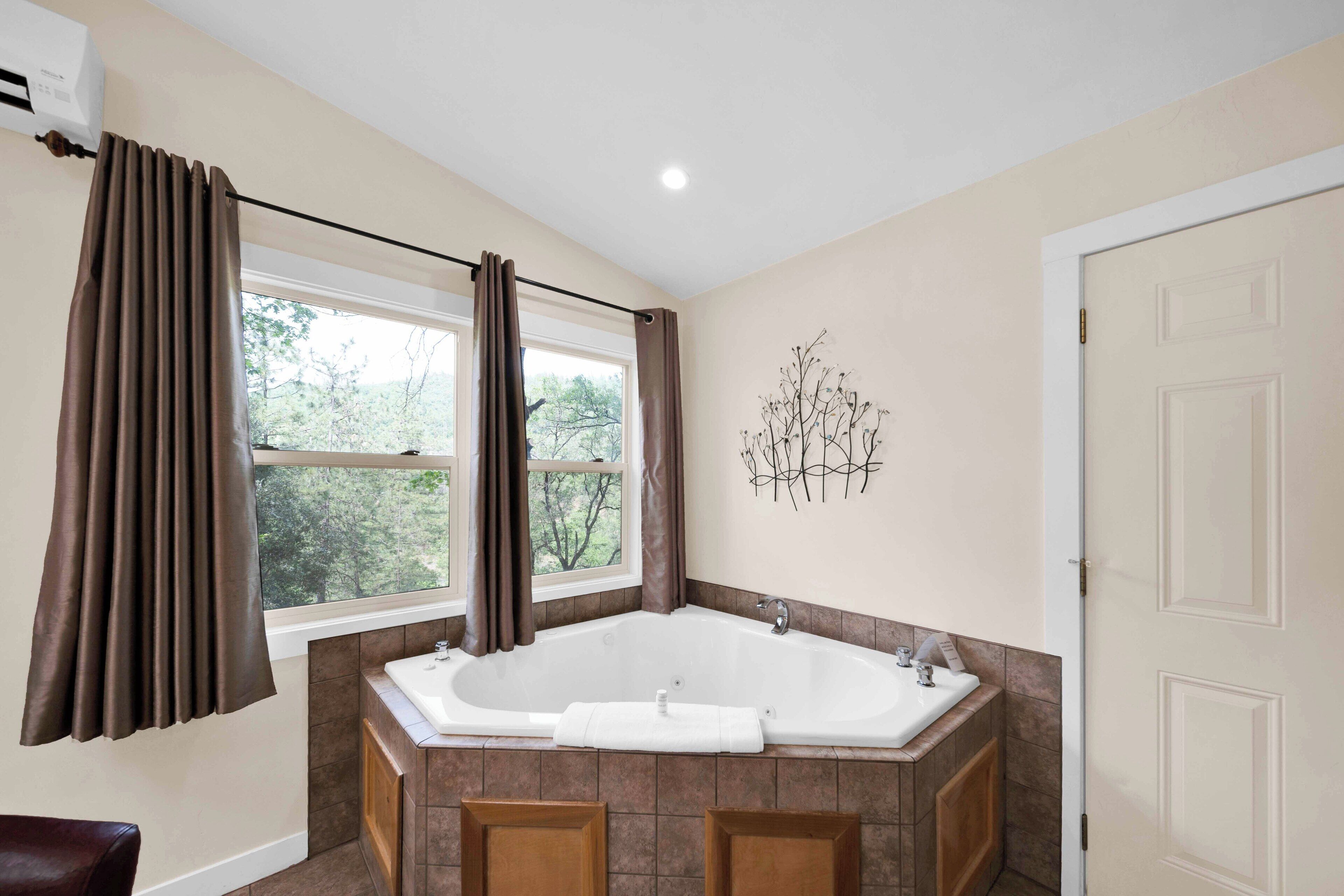 A bathroom with a large bathtub, a window with curtains, a sink, and a door.