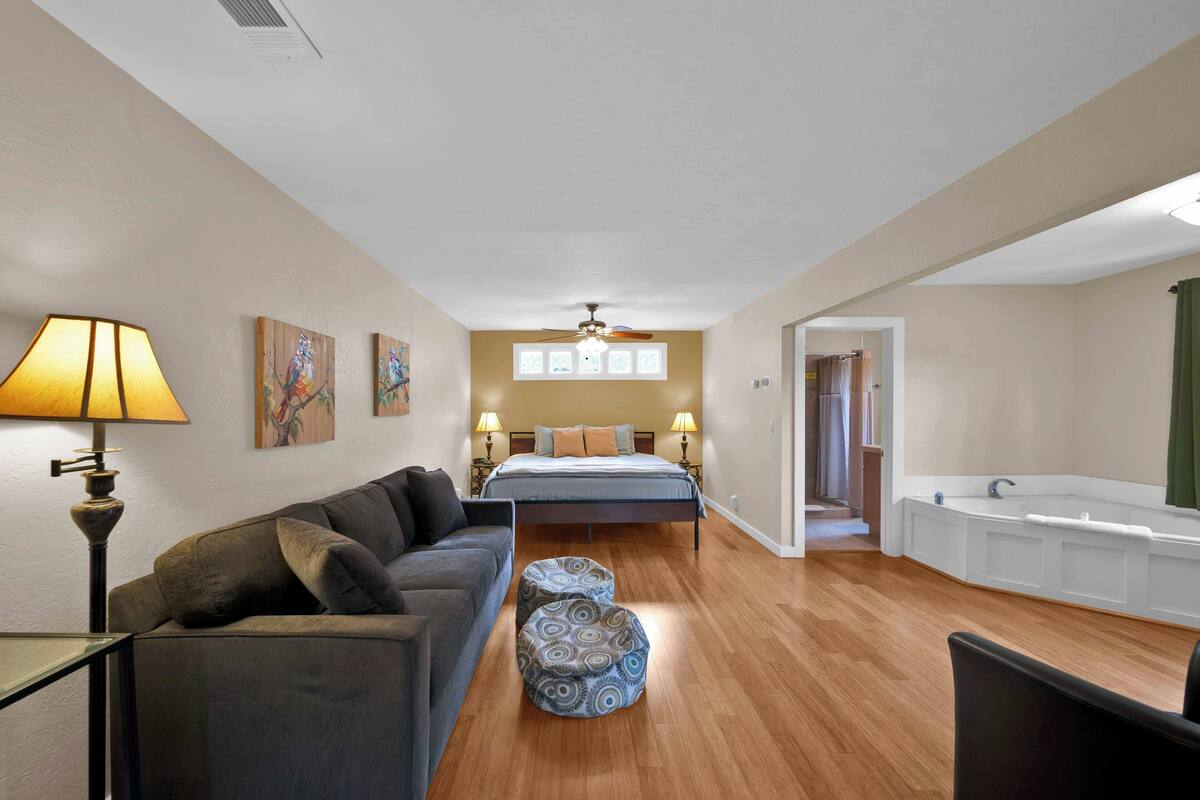 A bedroom with a bed, bedside lamps, a nightstand, a sofa, a patterned ottoman, a ceiling fan, and a bathtub.