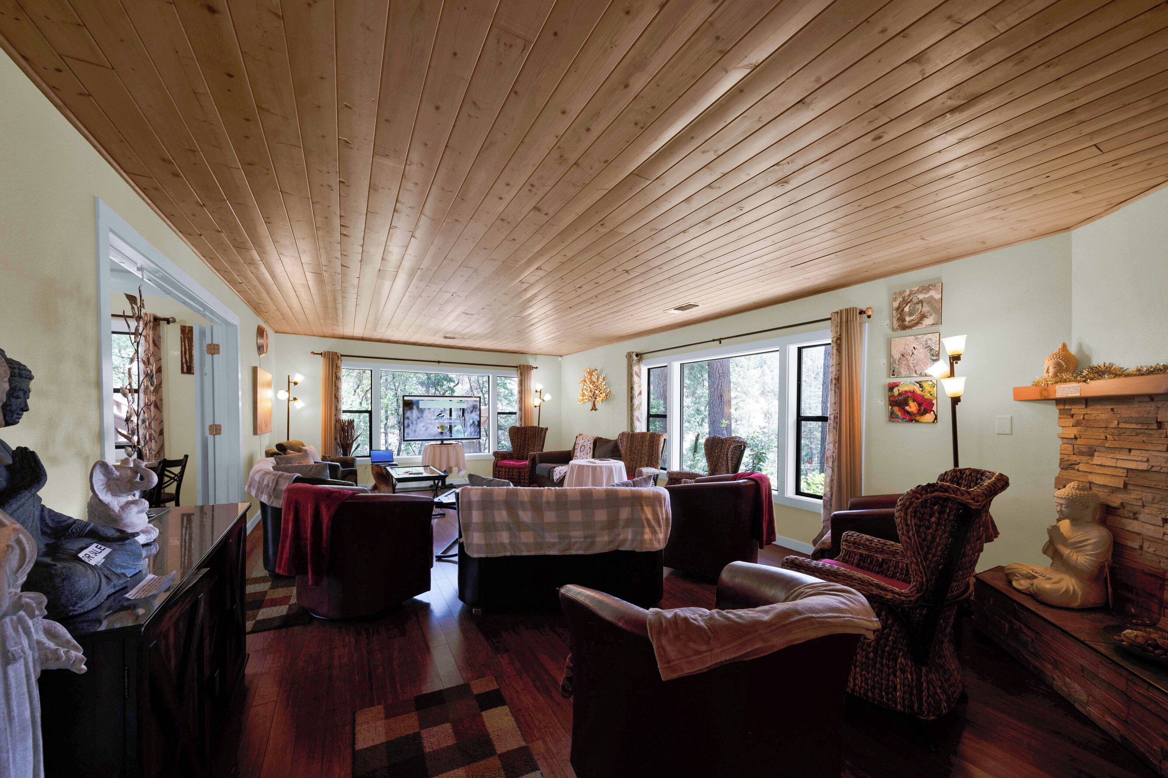 A spacious room with wooden ceiling, multiple seating areas, a fireplace, and large windows providing natural light.