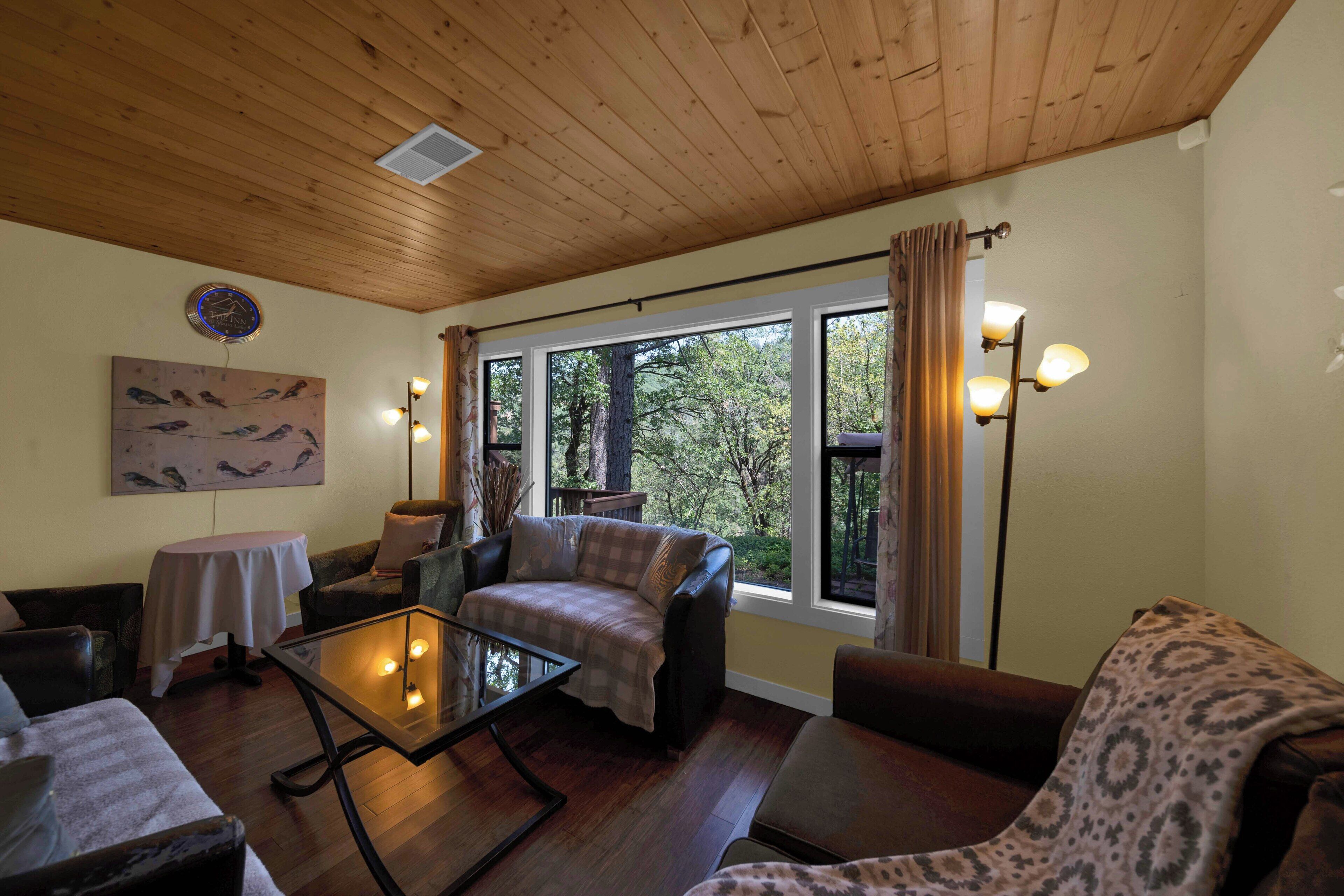 A cozy living room with a wooden ceiling, a large window with curtains, a glass coffee table, and a painting of birds on the wall.