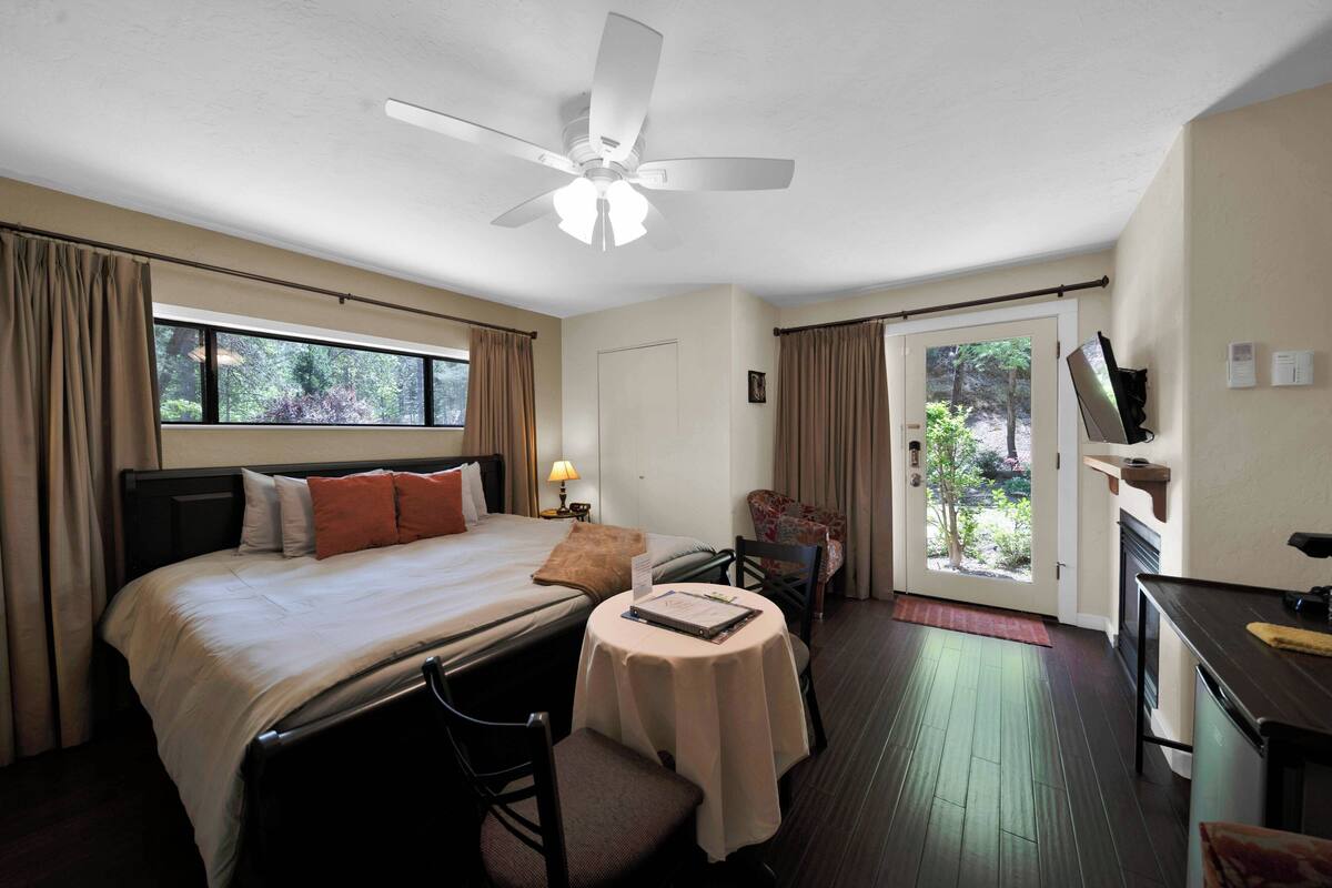 A hotel room with a bed, a desk, a chair, a small table, a ceiling fan, a TV, and a window with curtains.