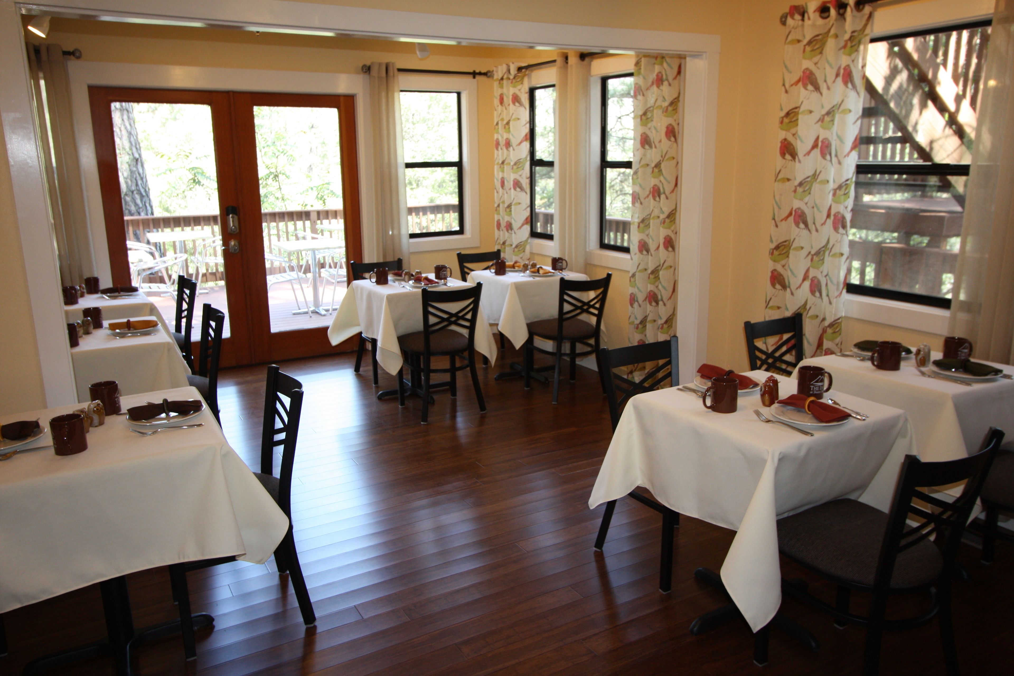 A restaurant with multiple tables and chairs, wooden flooring, and large windows with curtains.