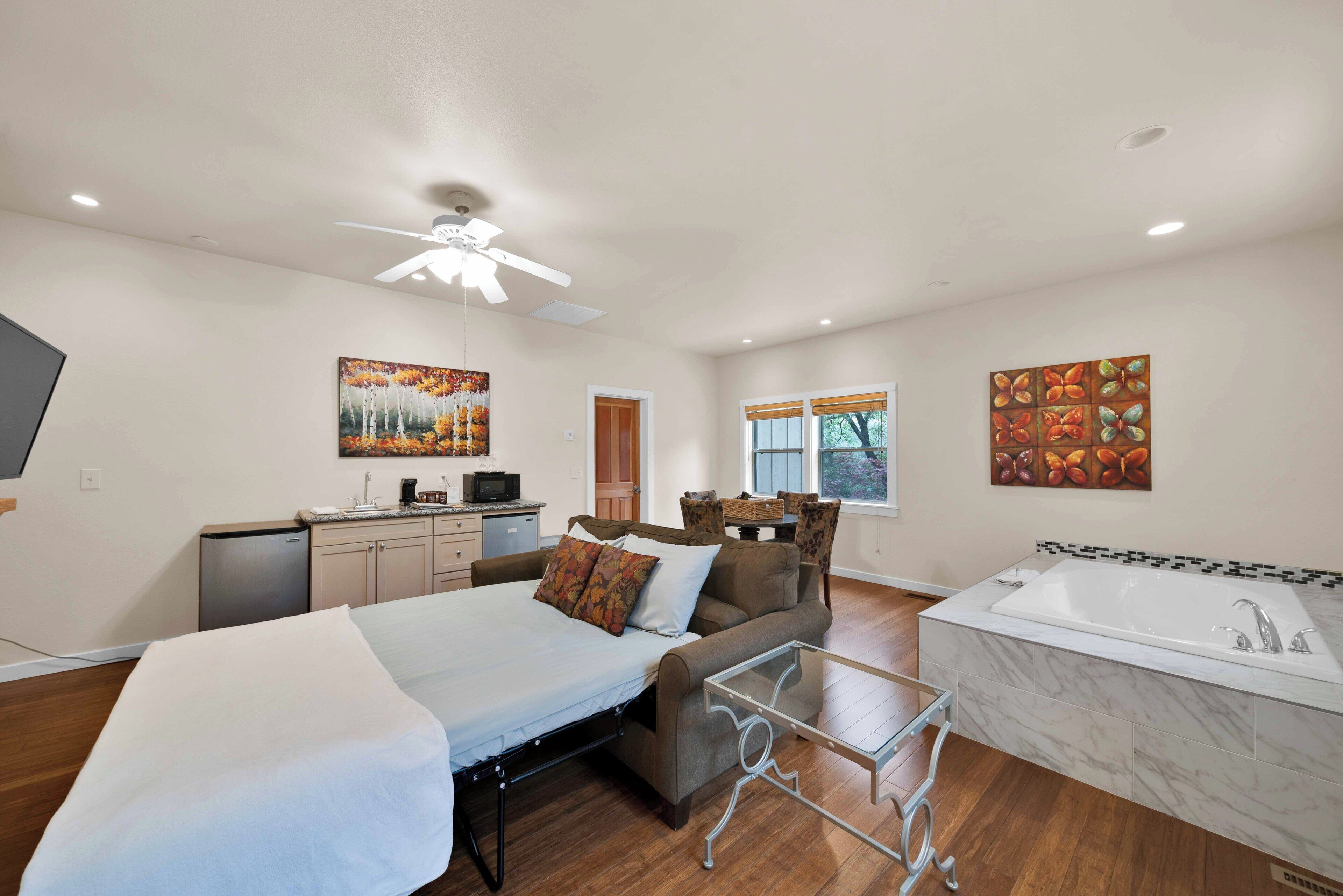 A hotel room with a bed, a sofa, a small table, a TV, a desk, a kitchenette, and a bathroom with a bathtub.