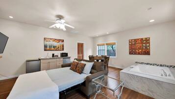 A hotel room with a bed, a sofa, a small table, a TV, a desk, a kitchenette, and a bathroom with a bathtub.