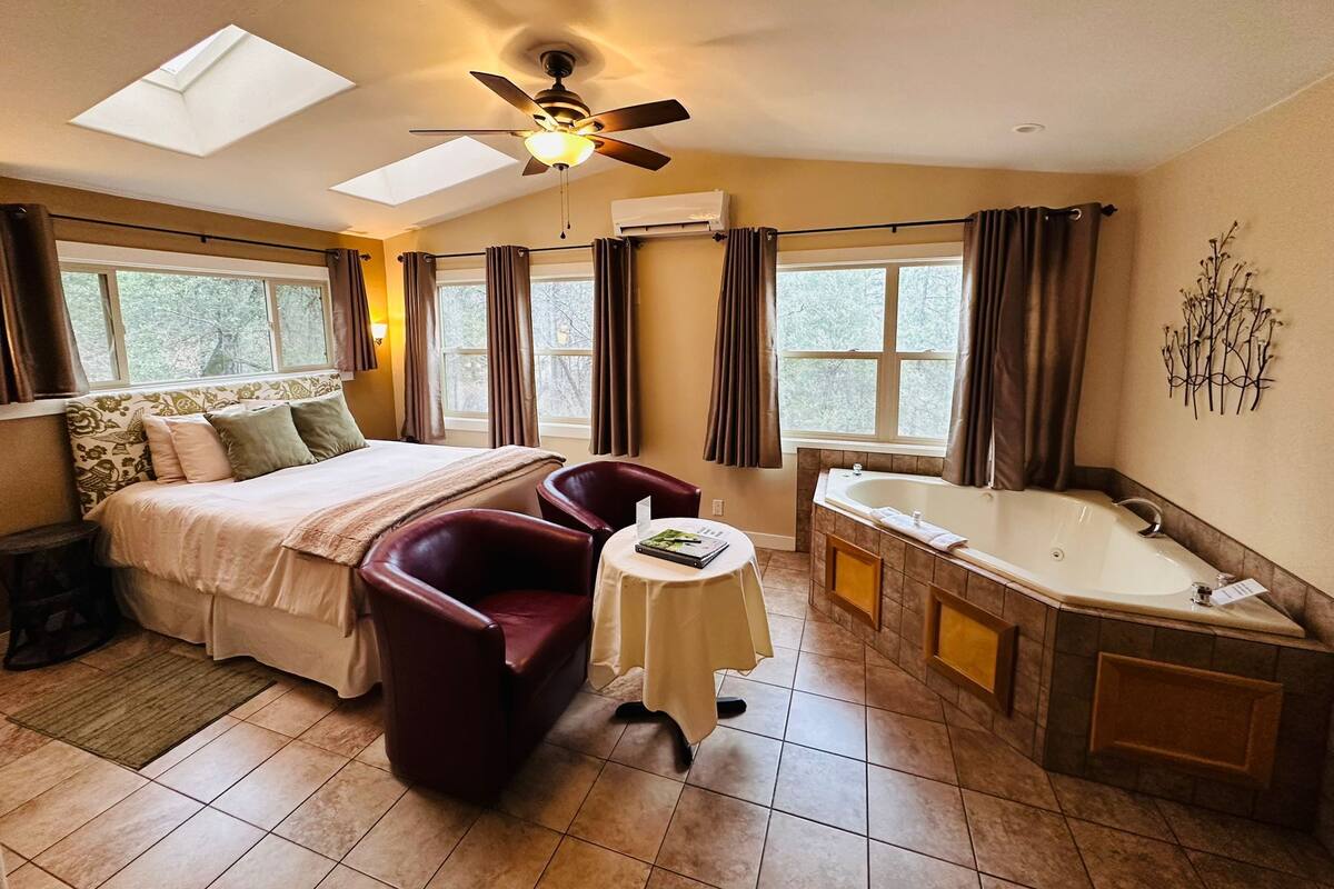 A bedroom with a bed, two chairs, a small table, a ceiling fan, and a bathroom with a bathtub and sink.