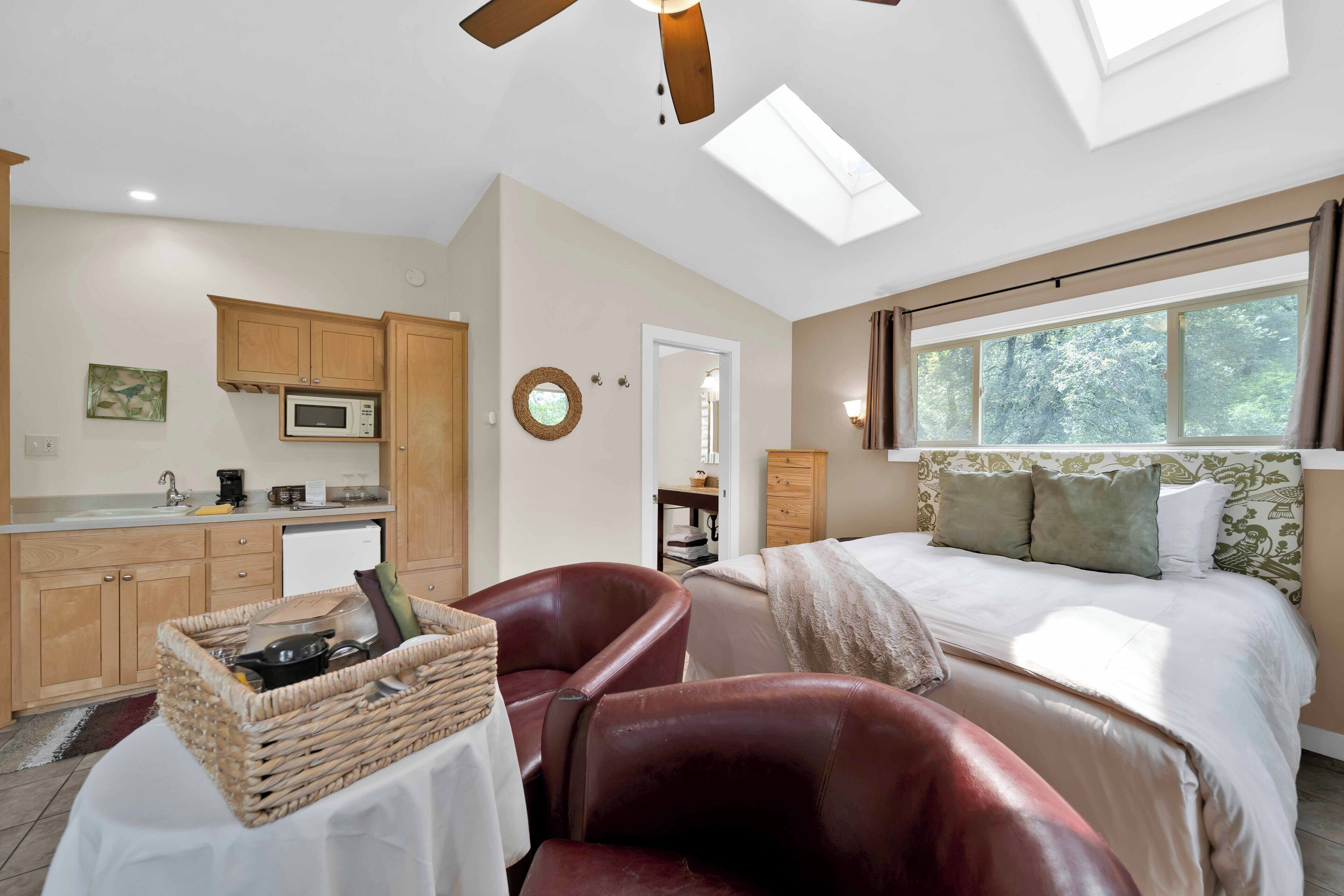 A bedroom with a bed, a desk, a chair, a window, and a kitchen area with cabinets and a microwave.