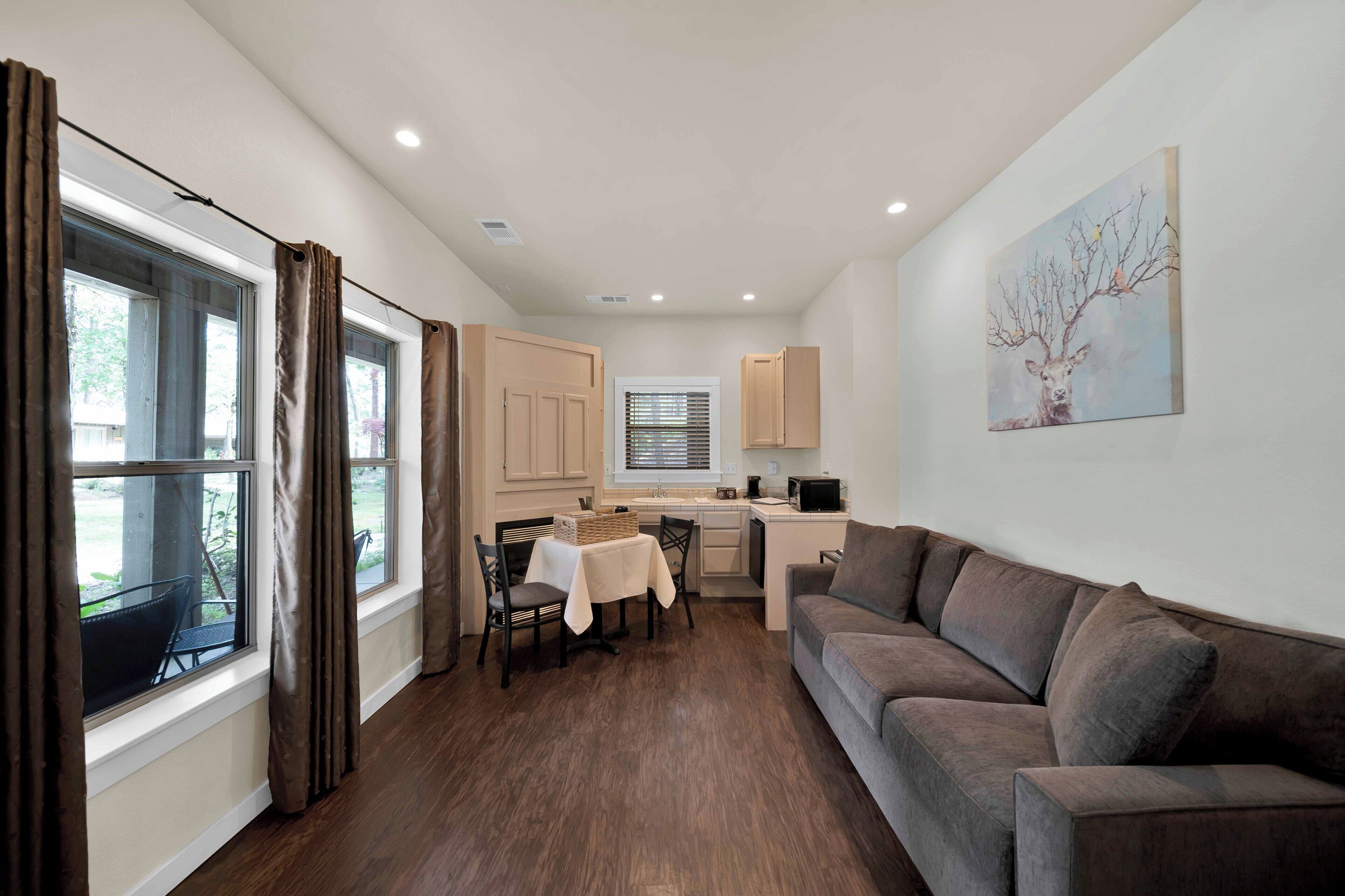 A modern living room with a sofa, a dining table with chairs, a kitchen area, and a large window with curtains.