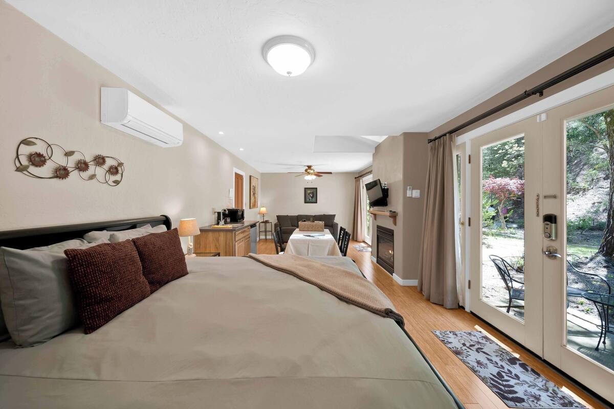 A bedroom with a large bed, a ceiling fan, a fireplace, a dining area, and a sliding glass door to the outside.