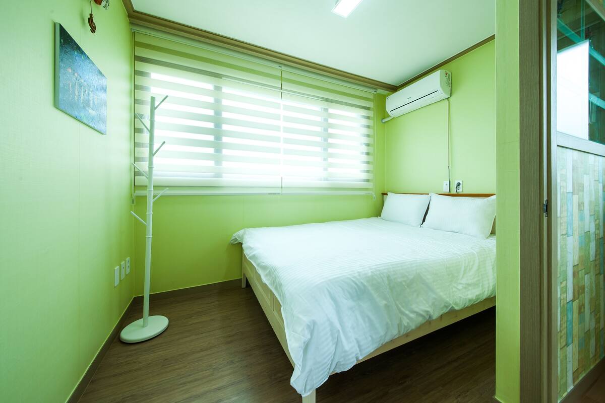 A room with a bed, a standing fan, a window with blinds, and an air conditioning unit.