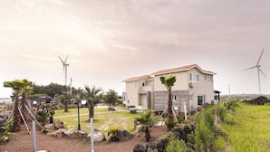 Une maison moderne à deux étages, avec un toit recouvert de tuiles rouges, entourée d’un jardin aménagé avec des palmiers et des rochers.