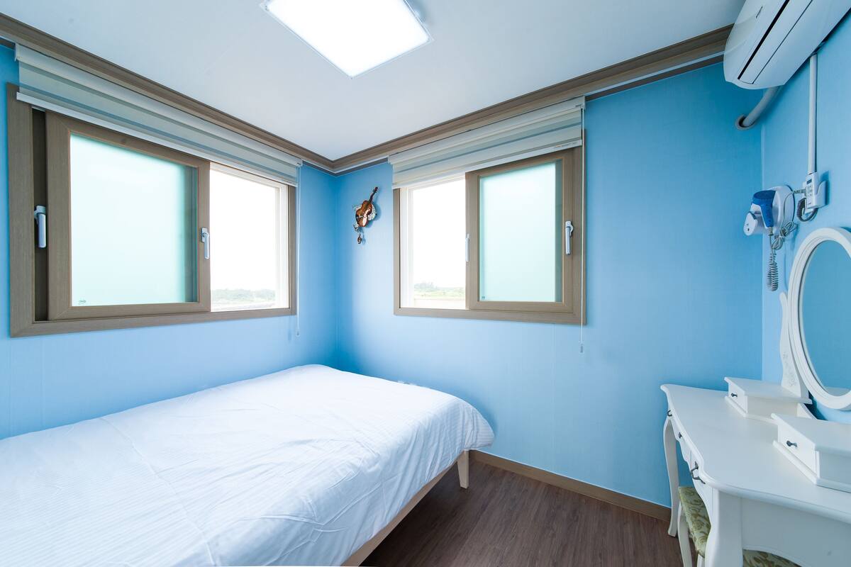 A bedroom with a bed, two windows, a vanity with a mirror, and an air conditioning unit.