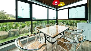 Un espace repas avec une table en bois et des chaises en métal, de grandes fenêtres offrant une vue sur les éoliennes, et un plafond orné de lampes suspendues.
