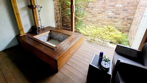 A wooden bathtub in a covered outdoor area with a brick wall and greenery in the background. - Positano in Jeju (Seogwipo)