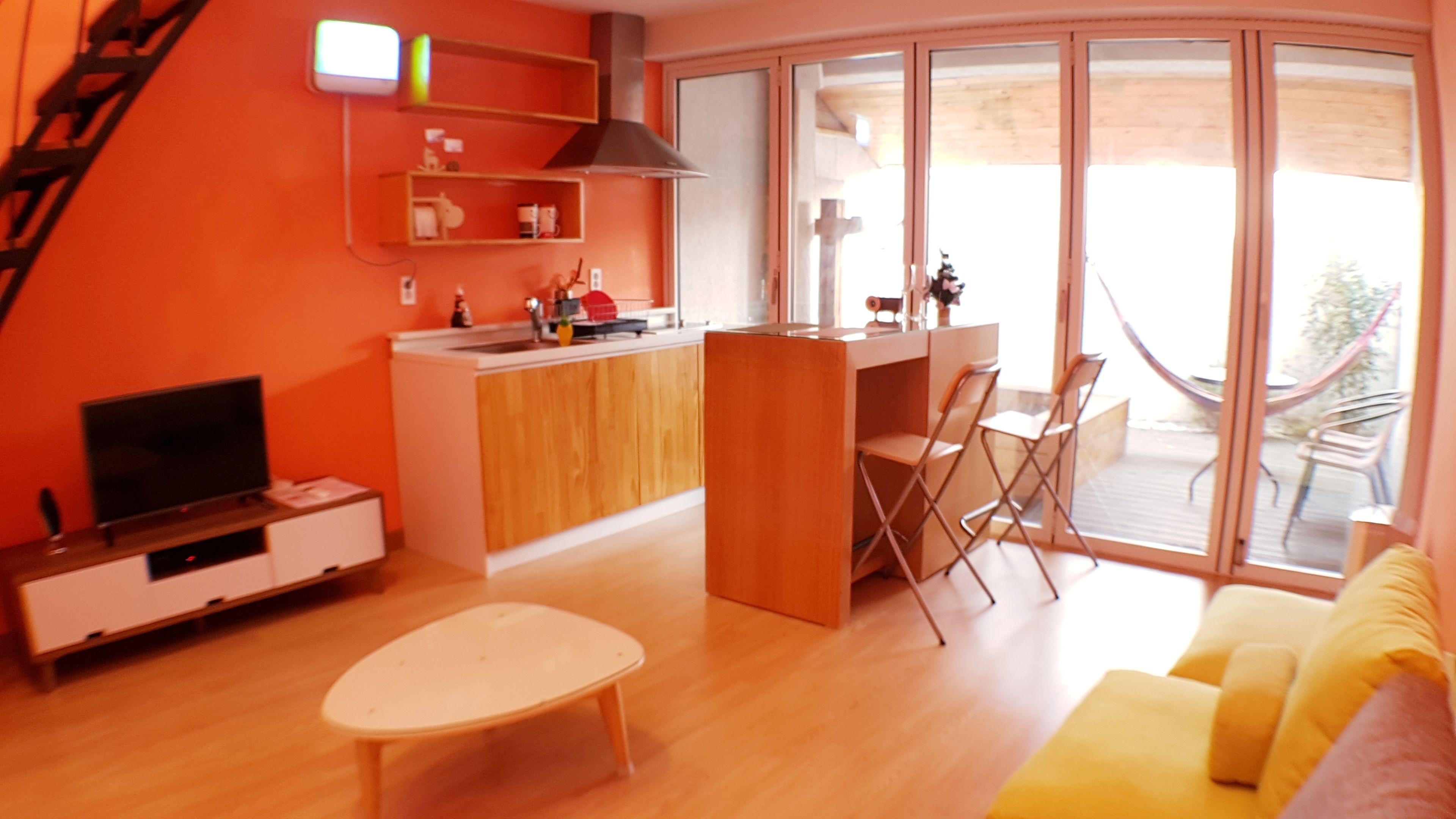 A modern living room with a TV, a small table, and a sofa. There is a kitchen area with a sink and cabinets, and a balcony with chairs.