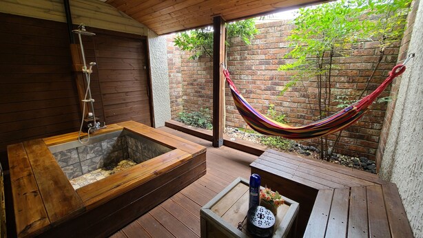 A wooden deck with a hammock, a wooden bathtub, and a small table with a bottle and flowers. - Positano in Jeju (Seogwipo)