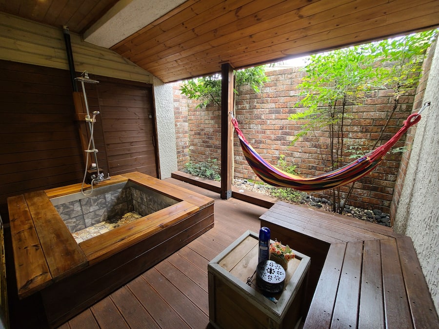 A wooden deck with a hammock, a wooden bathtub, and a small table with a bottle and flowers.