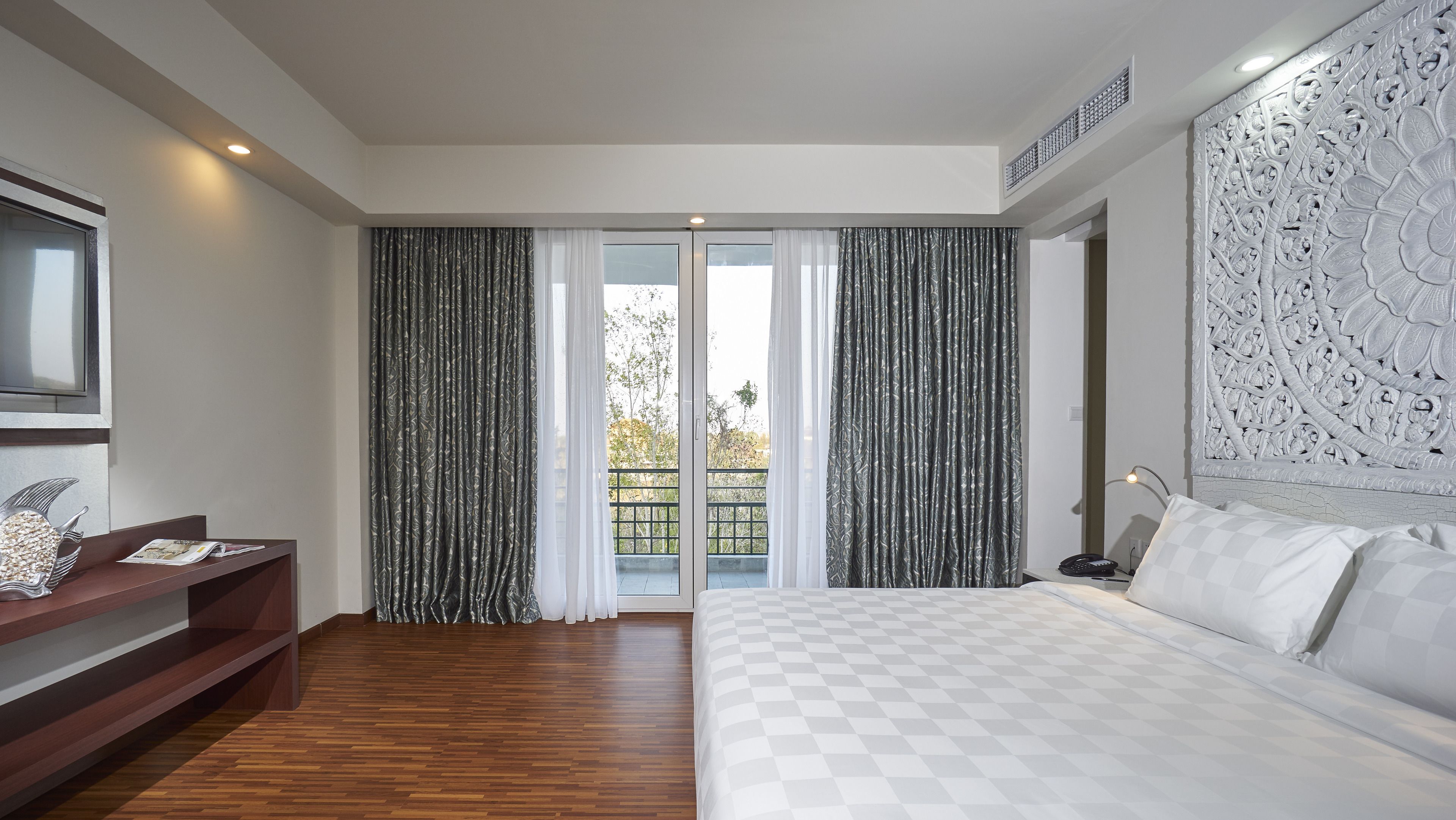 Une chambre d’hôtel moderne avec un grand lit, un bureau en bois et un balcon agrémenté de rideaux à motifs.