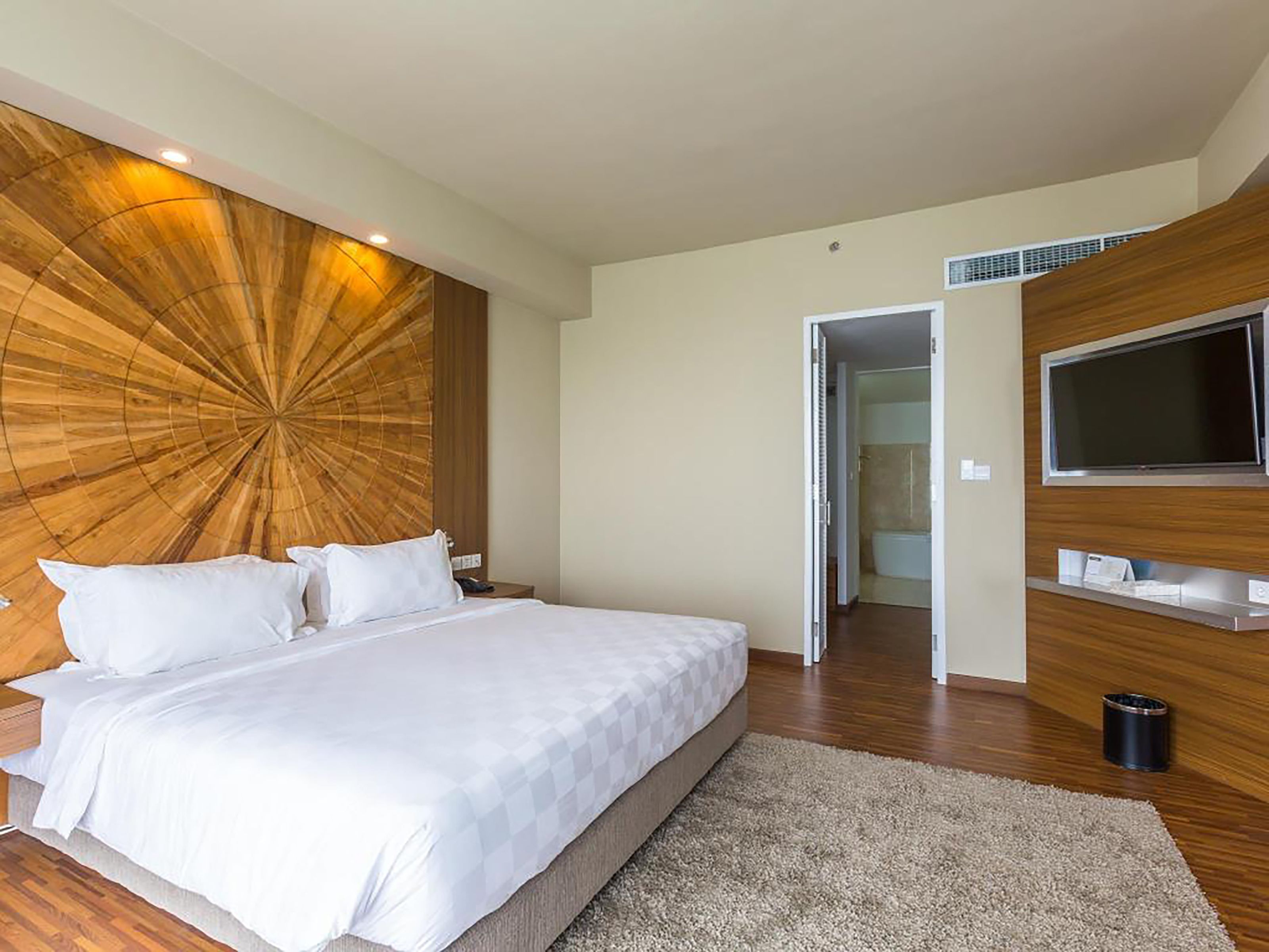 Une chambre d’hôtel moderne dotée d’un grand lit, d’un mur décoratif en bois, d’un téléviseur à écran plat et d’une salle de bain visible à travers une porte ouverte.