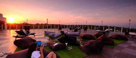 A rooftop lounge area with bean bags and lounge chairs, a sunset view, and a tiled floor.