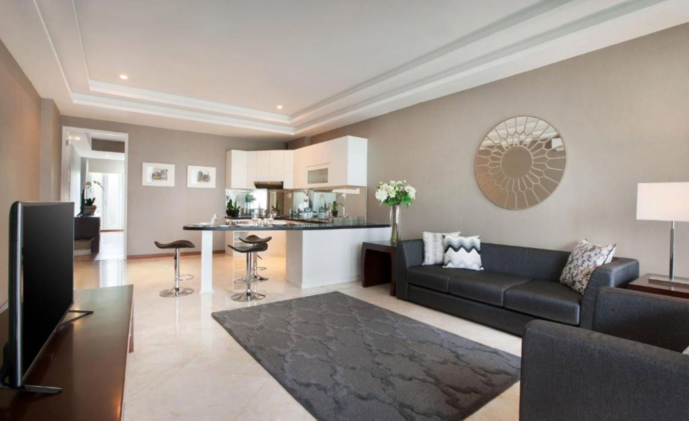 A modern living room with a dark grey sofa, a flat-screen TV, and a coffee table. There is a kitchen in the background with white cabinets and a central island.