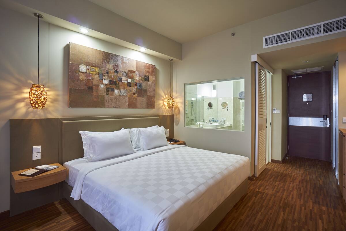 Une chambre d’hôtel avec un grand lit, une table de chevet, une œuvre d’art accrochée au mur et une salle de bain visible à travers une paroi en verre.