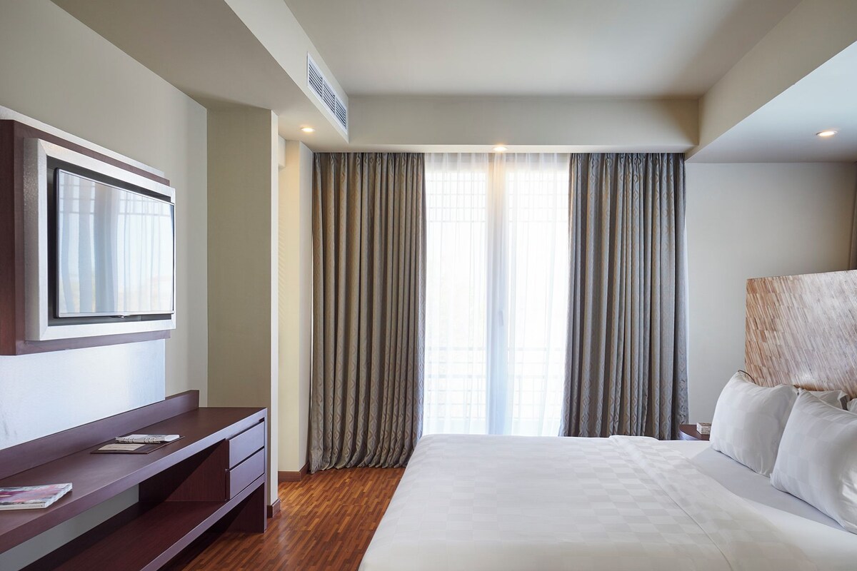 A hotel room with a bed, a desk, a TV, and large windows with curtains.