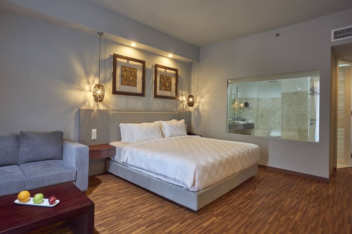 Une chambre à coucher moderne avec un grand lit, un canapé gris, une table basse en bois sur laquelle sont disposés des fruits, et un grand miroir.