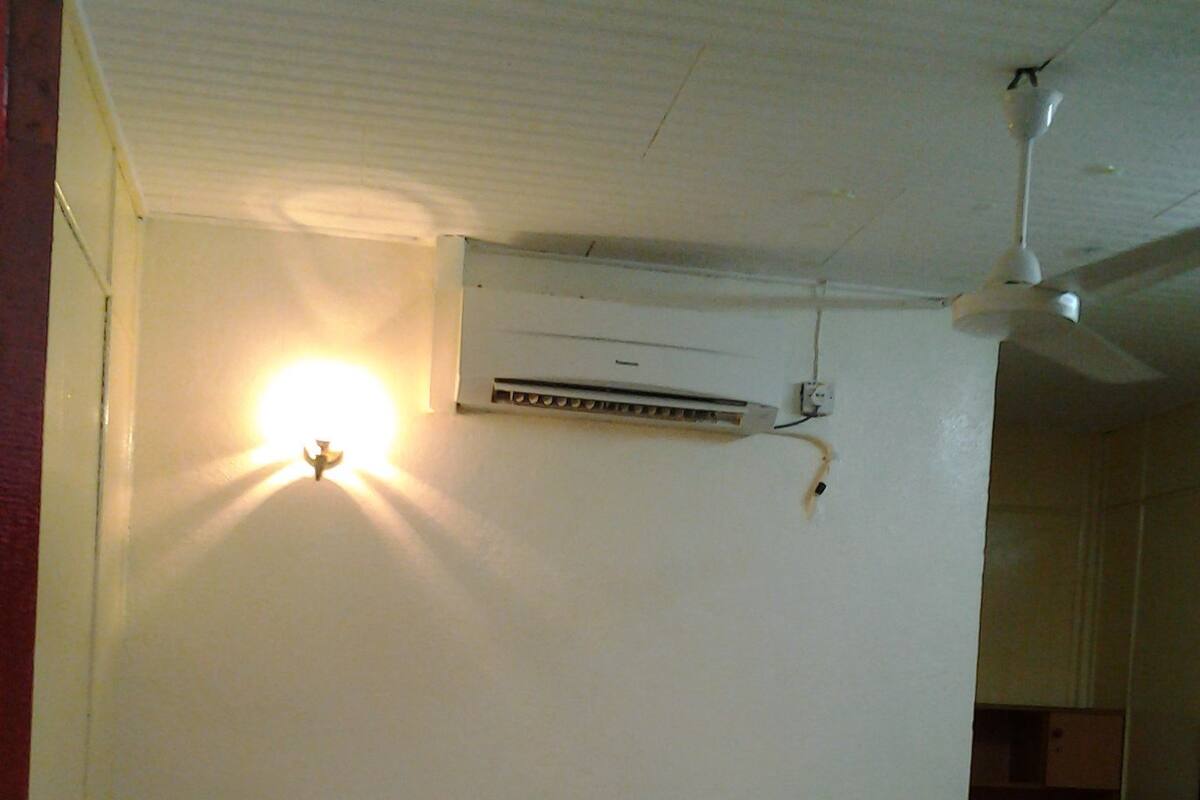A single bed with a white mattress and pillows, a wall-mounted air conditioner, and a ceiling fan.