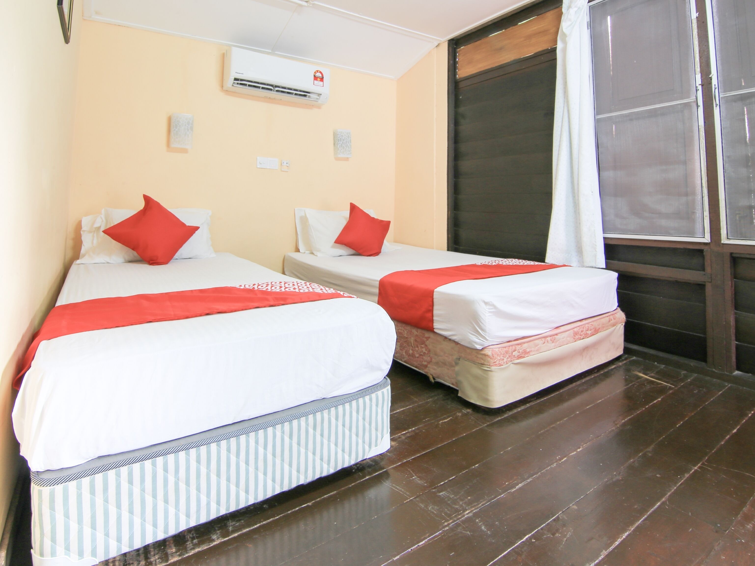 A hotel room with two beds, each with white bedding and red accents, a large window with a curtain, and a wooden floor.