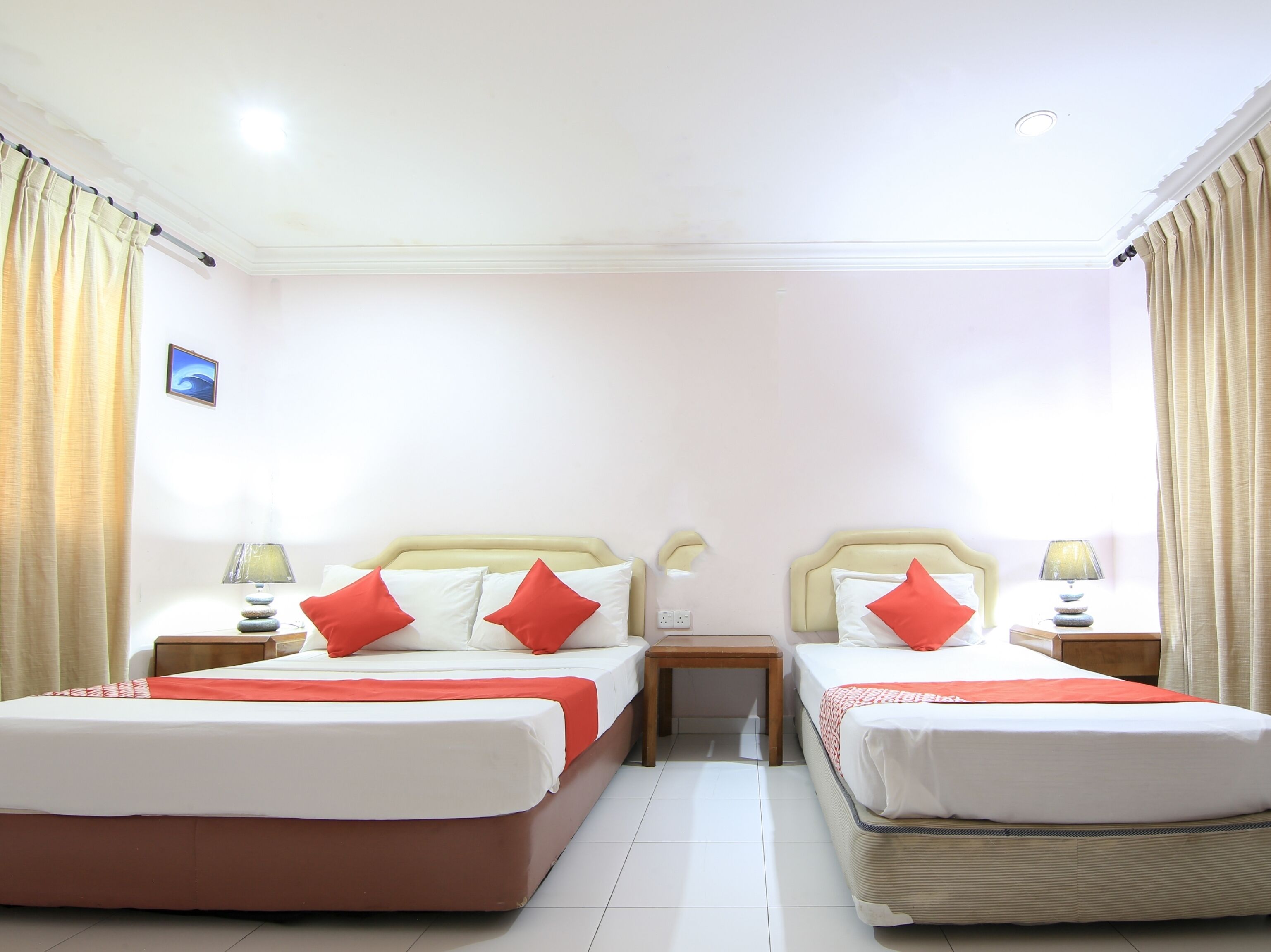 A hotel room with two beds, each with red pillows, a small table, and a nightstand with a lamp.
