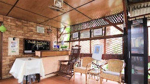 A restaurant with a brick wall, wooden ceiling, and tiled floor. There are wicker chairs, a reception desk, and a sign indicating breakfast and dinner times. - Hotel O Ombak Inn Chalet (Pangkor Island)
