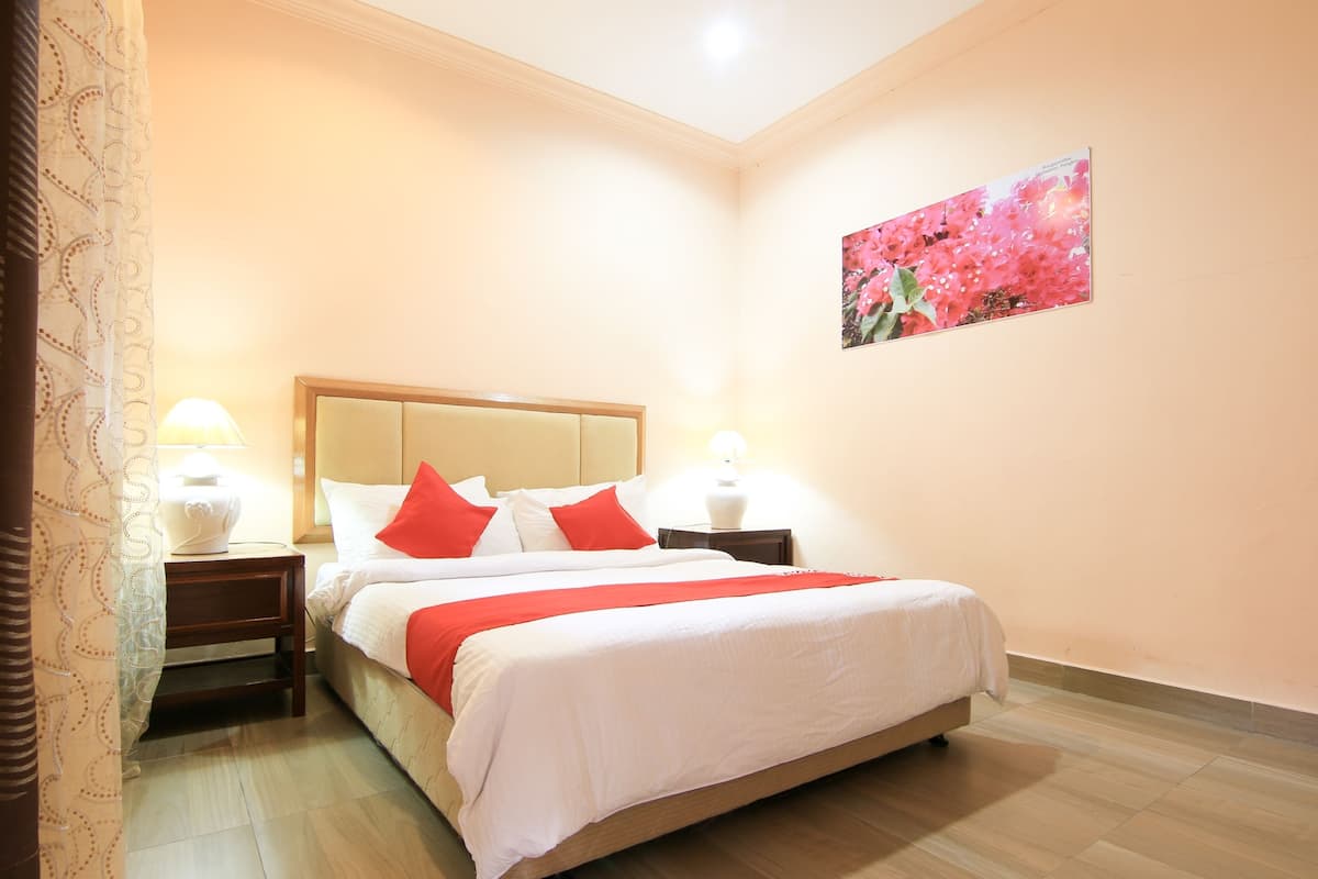 A hotel room with a bed, two bedside tables, a nightstand, a lamp, a curtain, and a floral painting on the wall.