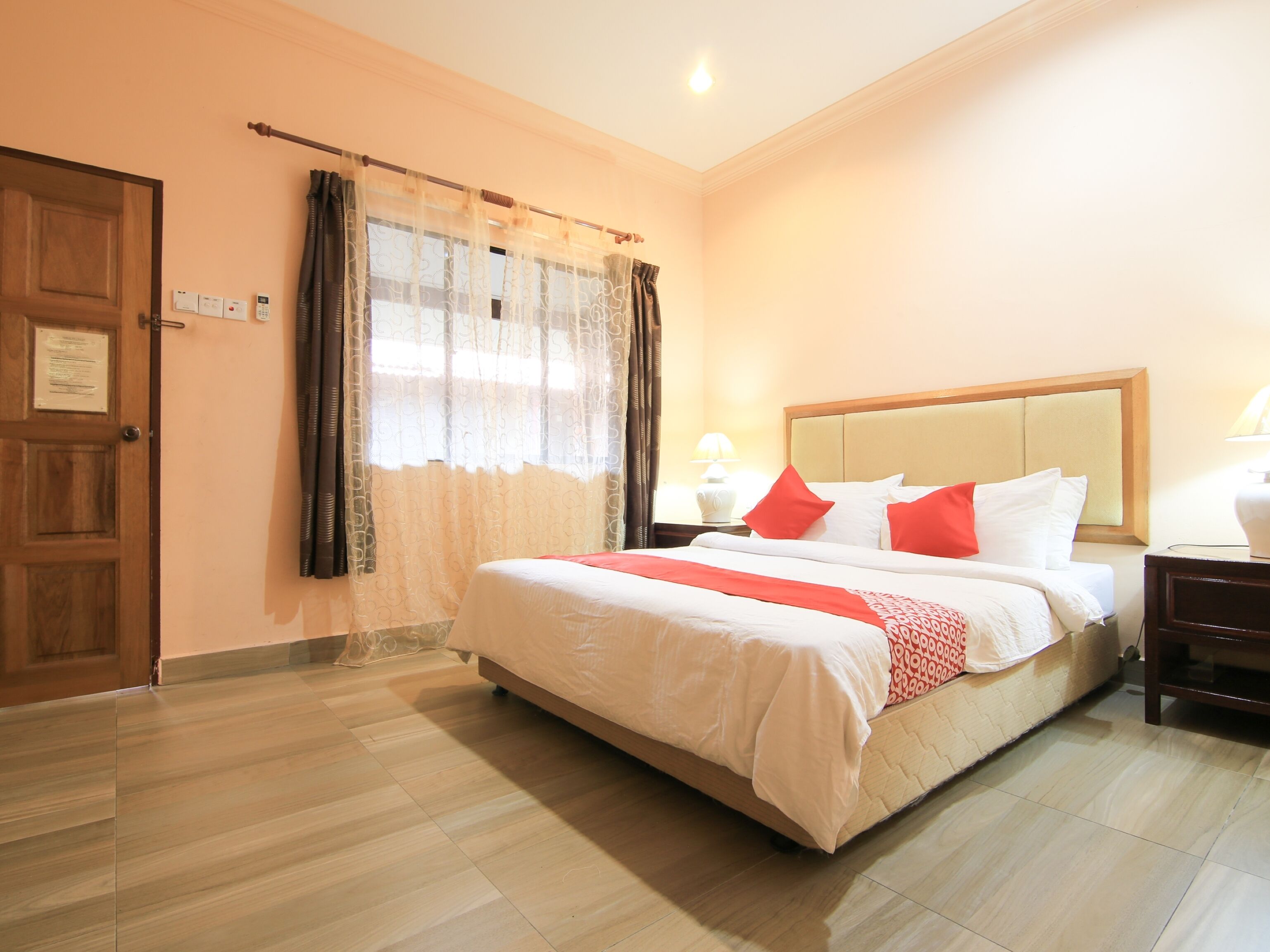 A hotel room with a bed, curtains, a wooden door, and a nightstand with a lamp.