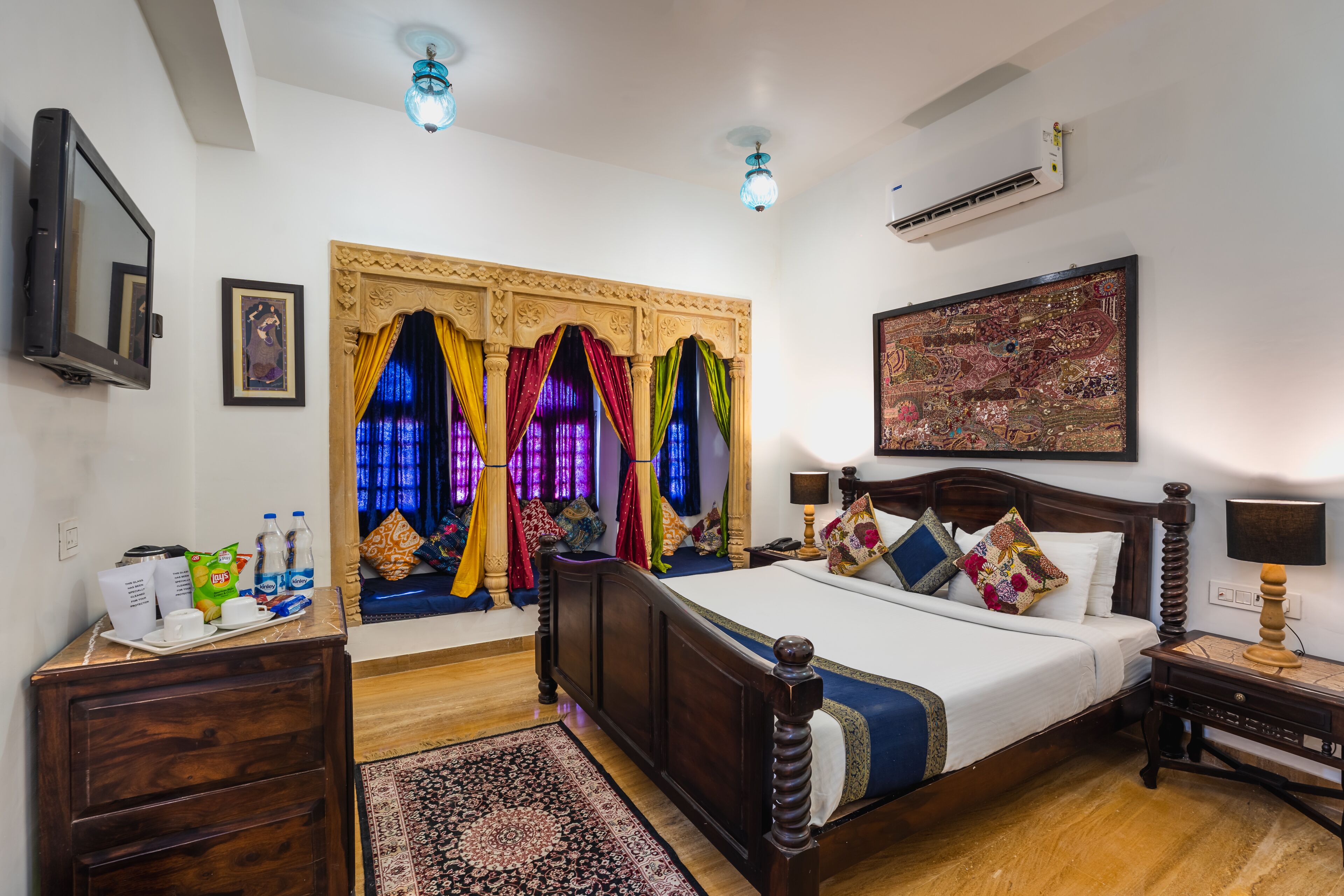 A hotel room with a wooden bed, a desk, a chair, a TV, a window with colorful curtains, and a wall-mounted air conditioner.