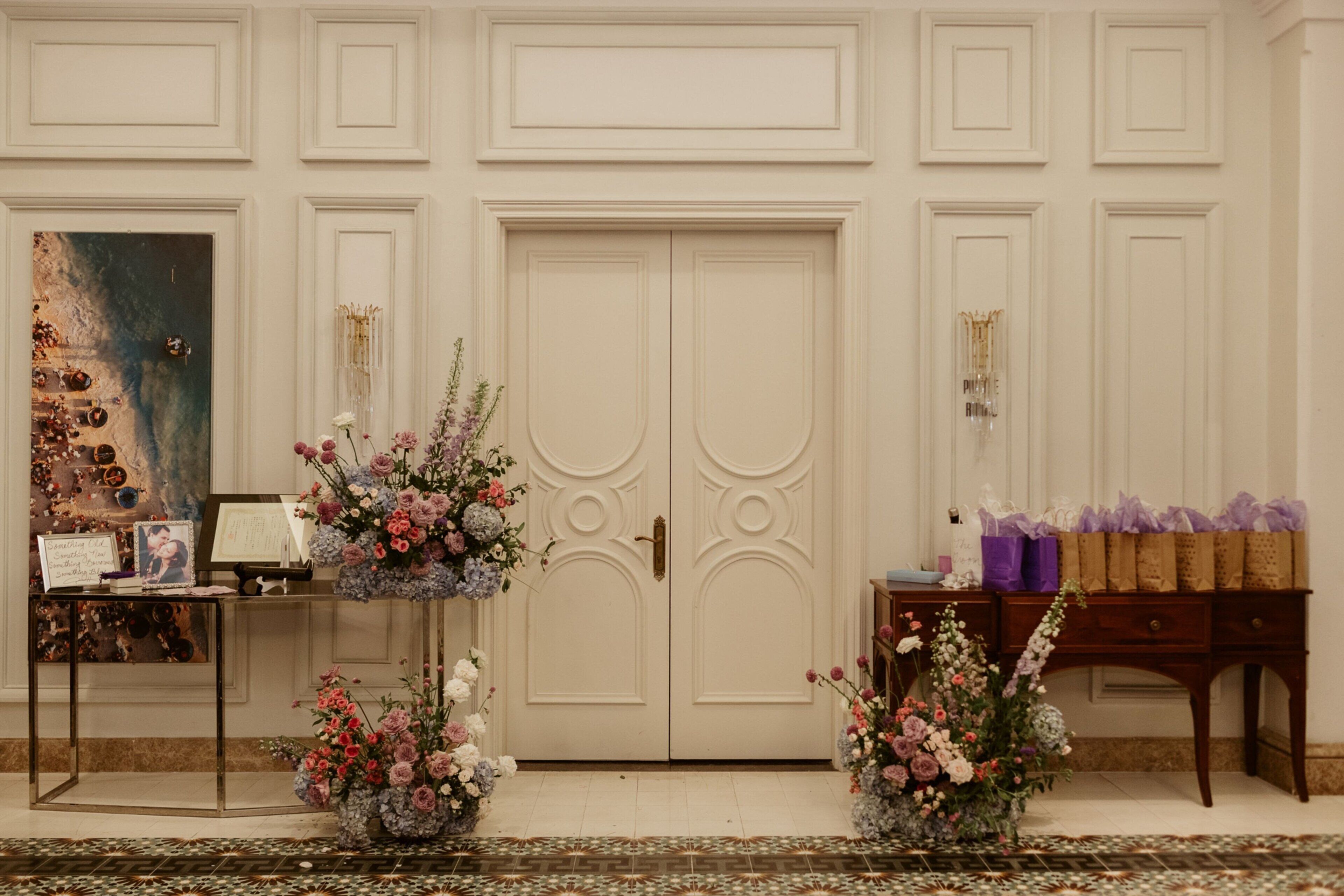 A room with a large white door, a small table with a vase of flowers, and a wooden side table with purple bags.