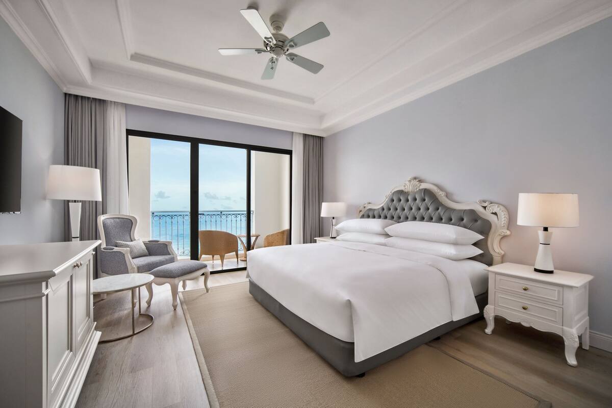 Een hotelkamer met een groot bed, een plafondventilator, uitzicht op de oceaan, een zithoek met een stoel en een tafel, en een dressoir.