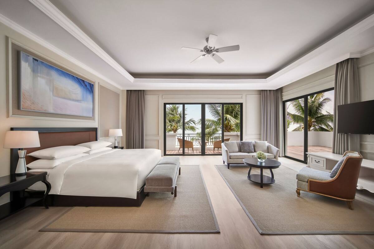 Een ruime hotelkamer met een groot bed, een zithoek met een salontafel, en een balkon met uitzicht op palmbomen.