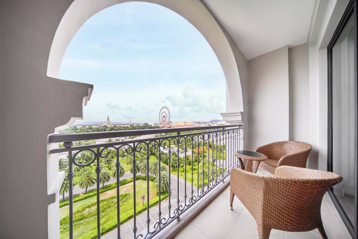 Een balkon met rieten meubilair, uitzicht op een park en een reuzenrad in de verte.