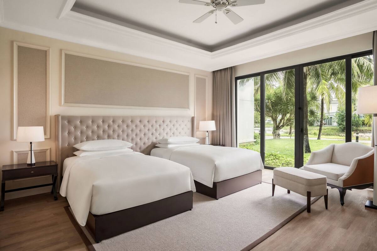 Hotelkamer met twee bedden, een zithoek en grote ramen met uitzicht op palmbomen.